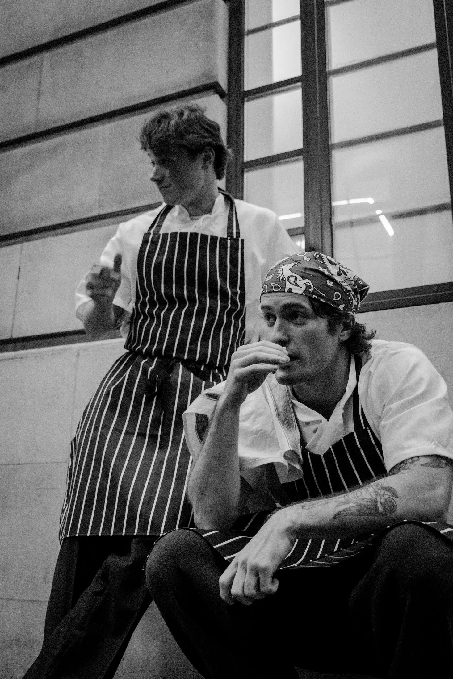 Two chefs in white jackets, one leaning on a counter and looking down.