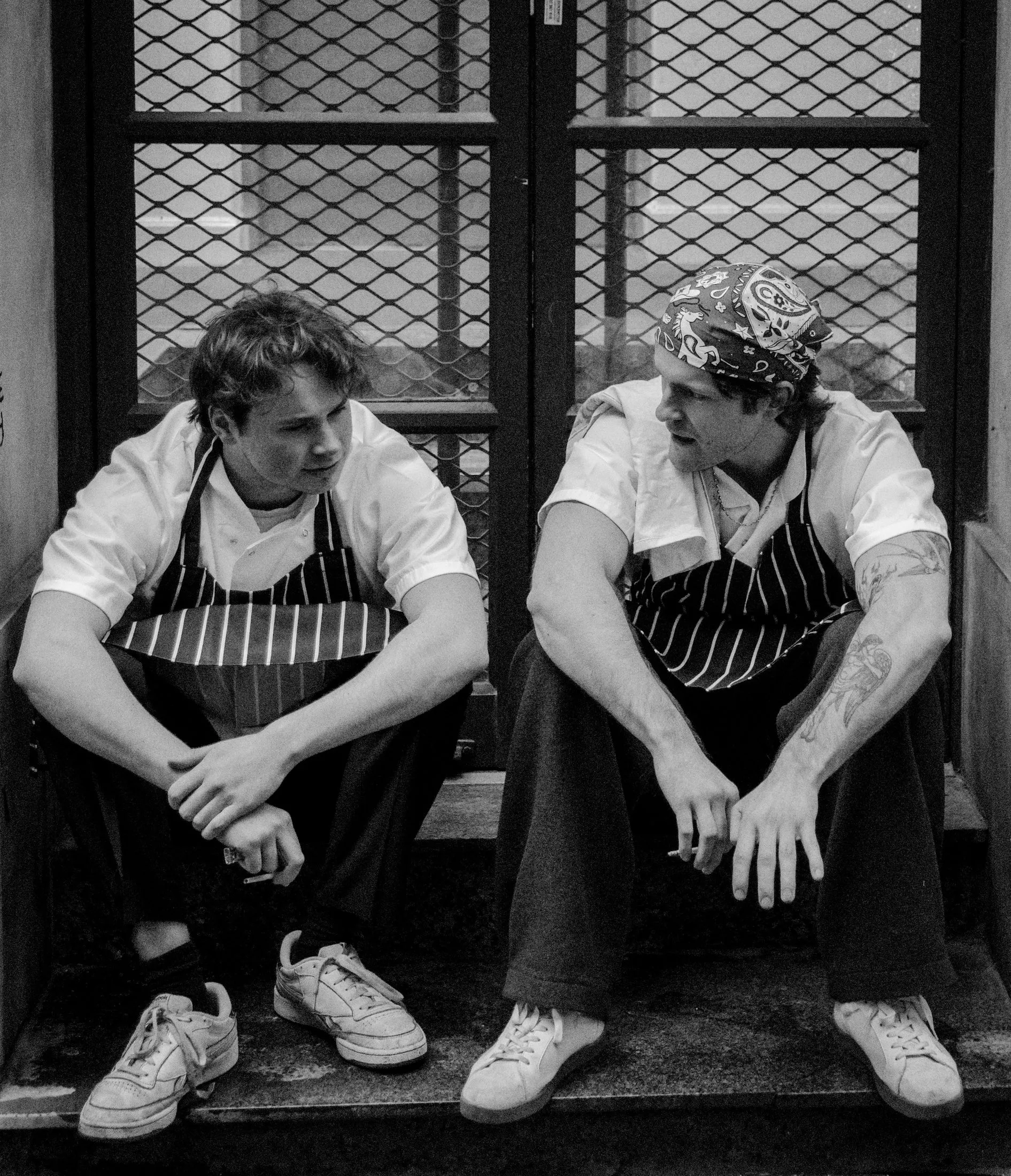 Two chefs in white jackets stand together in a kitchen, one turned partly away.
