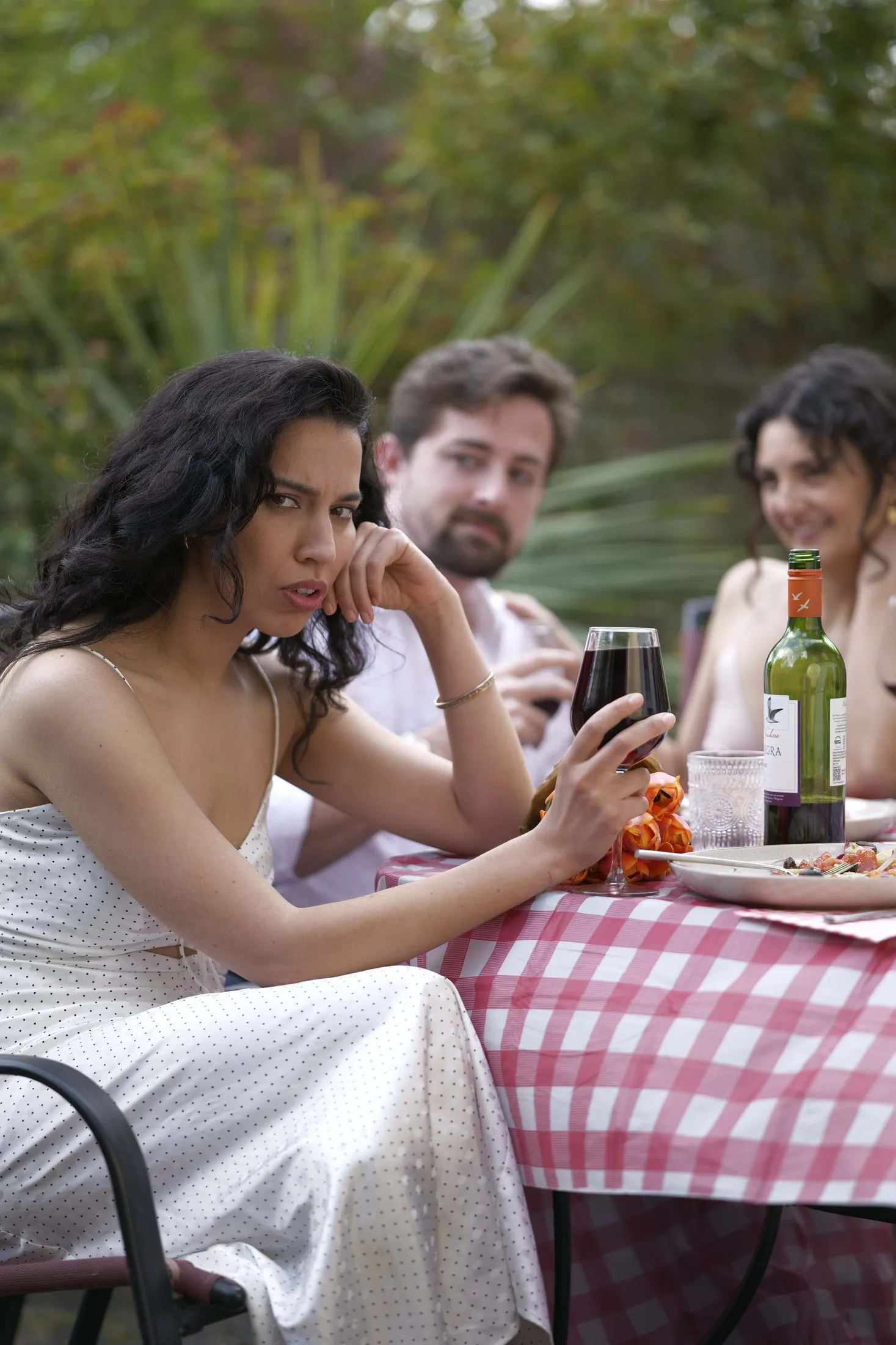 Two women at a dinner table, one in a white blouse and one in black, with wine glasses, plates and a red cloth.