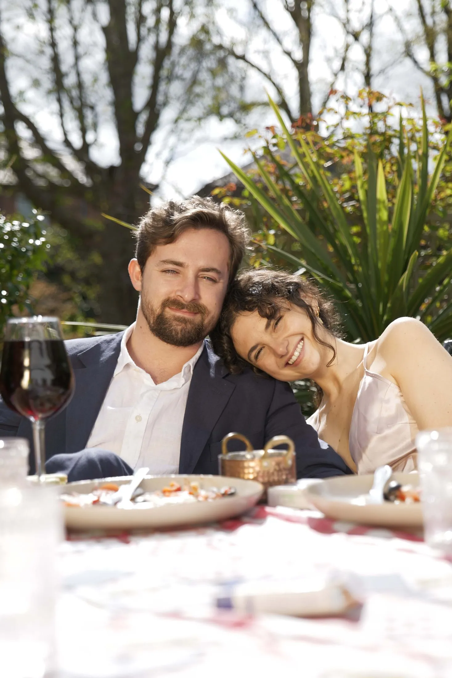A hand holds a glass of red wine above a set dinner table.